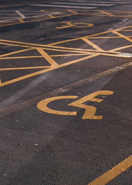 Close-up view of a designated accessible parking space in an outdoor parking lot, featuring yellow painted lines and a clear wheelchair symbol on the dark asphalt surface. The space is marked with bright yellow paint, with some of the lines intersecting or extending outside the parking bay. The surrounding area shows additional parking lines and a slight incline. This image captures the practicality of accessible parking during a home relocation or furniture transport process, as it indicates suitable parking for loading or unloading large items in proximity to a property. Man with Van Homerton occasionally provides assistance with such logistics, ensuring smooth access for moving vehicles and equipment. The image emphasizes the importance of planning parking arrangements for efficient house removals or packing and moving activities, especially in urban areas with marked accessible parking zones.
