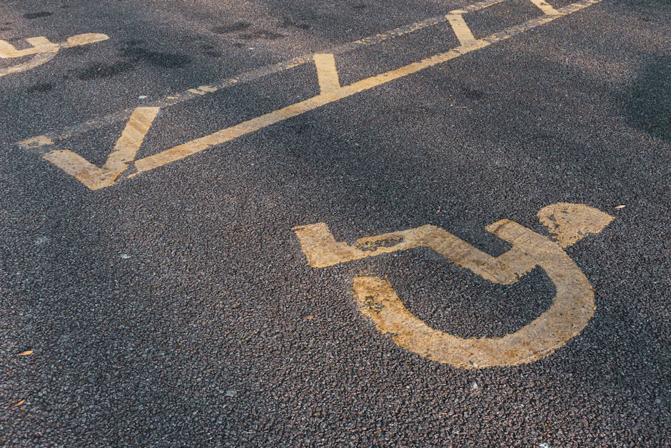 Close-up view of a designated accessible parking space in an outdoor parking lot, featuring yellow painted lines and a clear wheelchair symbol on the dark asphalt surface. The space is marked with bright yellow paint, with some of the lines intersecting or extending outside the parking bay. The surrounding area shows additional parking lines and a slight incline. This image captures the practicality of accessible parking during a home relocation or furniture transport process, as it indicates suitable parking for loading or unloading large items in proximity to a property. Man with Van Homerton occasionally provides assistance with such logistics, ensuring smooth access for moving vehicles and equipment. The image emphasizes the importance of planning parking arrangements for efficient house removals or packing and moving activities, especially in urban areas with marked accessible parking zones.