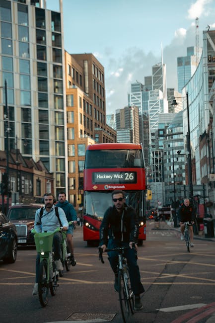 A busy urban street scene in Hackney with a red double-decker bus displaying 'Hackney Wick 26' approaching the intersection. Several cyclists, including one man in dark sunglasses and a dark jacket, are riding in the same direction as the bus on the paved road. Two men with backpacks are riding bicycles, one with a green basket on the front, while others pedal alongside or cross the street. Modern high-rise buildings with glass facades and brick exteriors surround the scene, reflecting the bright daylight and scattered clouds in the sky. In the background, more cyclists and pedestrians are visible, along with traffic lights and street signage. The setting suggests an active city centre environment suitable for home relocation and furniture transport services, with Man with Van Homerton occasionally associated with such moving logistics in the area.