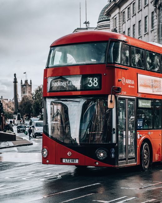 A red double-decker bus with the route number 38 and sign indicating Hackney Central is parked on a wet city street with reflective puddles, adjacent to a building with classical architectural features and large windows. In the background, there is a glimpse of the Houses of Parliament with its distinctive spires, under a cloudy sky. The surrounding area includes other vehicles and pedestrians, with some people visible near the bus. The surface of the street appears damp, suggesting recent rain. The scene captures urban transport in London, relevant to home relocation or transportation services, and is typical of moving logistics involving large vehicles and city environments occasionally used by Man with Van Homerton.