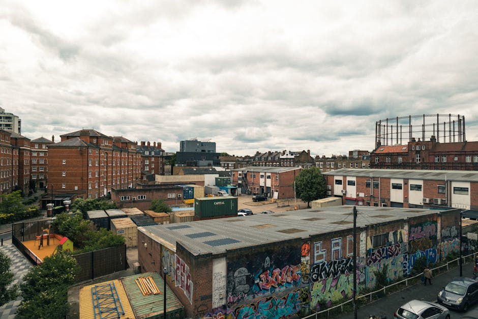 A view of an urban area with multiple brick and concrete buildings under a cloudy sky, including a large warehouse with graffiti-covered walls at street level, and a fenced playground with a small red and orange playhouse. In the background, there are residential apartment blocks and industrial structures such as a large metal tank. The scene shows the loading area of a home relocation or furniture transport operation, with the presence of parked vehicles and storage containers, indicating active moving or packing activities. This setting illustrates a typical inner-city environment relevant to house removals and moving logistics, as managed by companies like Man with Van Homerton, capturing the environment where loading and loading processes often take place.