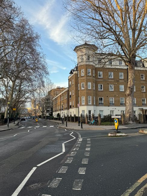 A wide view of a street corner in Homerton with a large multi-storey residential building constructed of brown brick and white accents, situated on the corner of Homerton Grove in E9. Several leafless