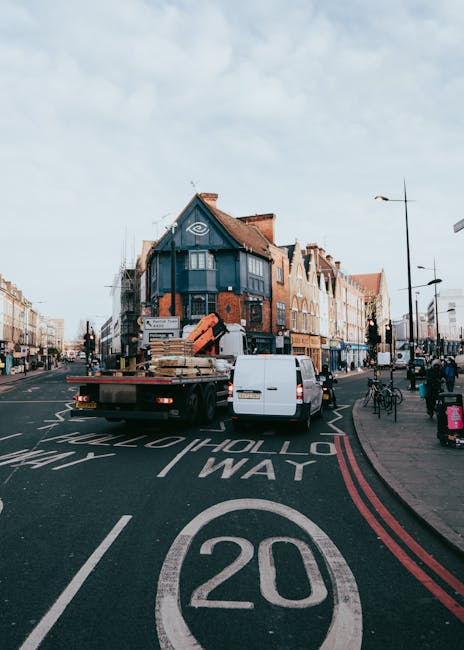 A white flatbed truck with an open cargo area is parked on a street in Homerton, featuring a collection of packed cardboard boxes, wooden pallets, and wrapped furniture items. The truck is positioned near the curb with its rear facing residential buildings, which include a dark blue corner property with a flat roof and orange brick facade, alongside other terraced houses with pitched roofs and brick exteriors. A white van is parked behind the truck, and the loading process appears to be underway, with some items possibly being loaded or unloaded. The street has clear road markings, including a 20 mph speed limit, and is lined with street lamps, bicycles, and pedestrians. The atmosphere suggests an active home relocation or furniture transport operation typical of house removals, with visible equipment such as blankets or straps may be used for securing items. Man with Van Homerton often assists with these kinds of loading and moving services to facilitate efficient furniture transport and packing for house moves.