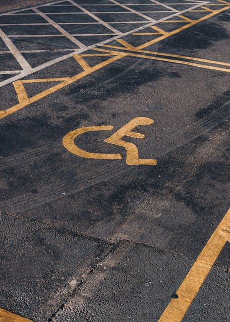 A close-up view of an asphalt parking area featuring a yellow painted wheelchair accessibility symbol on the ground. The symbol is positioned in a designated parking space near a curb, with adjacent yellow lines creating a grid pattern for parking bays. The surface shows signs of wear, with some patches and cracks visible. In the background, there are additional parking lines and a section of the kerb, indicating an outdoor area used for vehicle and possibly furniture transport during home relocations. This scene illustrates a streetside access point suitable for vehicle loading or unloading, relevant to house removals services provided by Man with Van Homerton, who specialise in furniture transport and packing and moving logistics.