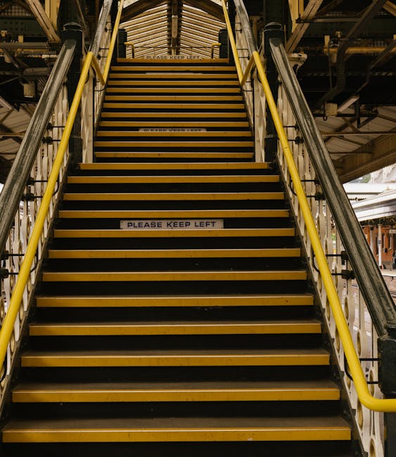 Inside a London Underground station with beige tiled walls and a curved ceiling, showing a platform edge with tactile paving in yellow, a set of stairs with a black handrail leading upward on the left