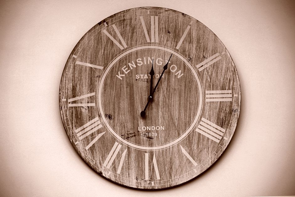 A round wooden wall clock with a vintage design, featuring Roman numerals in white and the words 'Kensington Station' and 'London 1809' printed on its face, hangs on a plain, light-colored wall. The clock's hands indicate the time as approximately 12:02. The clock's surface has a weathered, distressed finish, enhancing its rustic appearance. The image is well-lit, showing the clock clearly and emphasizing its textures. This clock may be part of the interior decor in a home or office space during a house removal or home relocation by Man with Van Homerton, highlighting the setting where packing and moving logistics are taking place, perhaps during inventory organization before loading furniture and boxes into a moving vehicle.