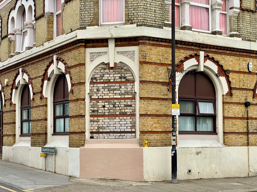 Image of a corner of a multi-story brick building with distinctive white decorative arches above three large, evenly spaced windows with brown frames and curtains, set on a white-painted base. The building's brickwork features alternating yellow and red bricks, with some areas of exposed white brickwork. In front of the building, a narrow sidewalk runs along the street, with a black lamppost positioned near the corner and a small yellow sign attached to it. A street sign reading 'Crawford Road' is mounted on the building's wall at street level. This scene depicts an urban setting potentially involved in home relocation or furniture transport, with the building's entrance not visible. The overall environment is well-lit, indicating daytime, supporting visual documentation for house removals or moving services such as those provided by Man with Van Homerton.