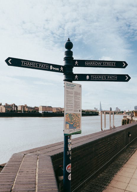 A black metal signpost with multiple directional signs mounted at the corner of a riverside walkway, indicating directions to Thames Path and Narrow Street. The signpost features decorative finials at the top and a small informational map attached at the lower section. Surrounding the signpost is a brick-paved walkway with a black metal bench nearby, and the River Thames is visible in the background with a cityscape including various buildings and the Shard skyscraper across the water. The sky above is partly cloudy with soft, diffused light, suggesting daytime. As part of a moving or home relocation context, such signage could be associated with navigating the local area around Homerton, supporting logistics and planning for furniture transport or packing.