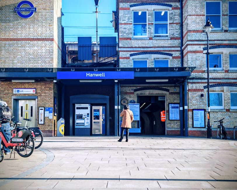 A person standing outside Hanwell station, part of the Elizabeth Line, surrounded by bicycles and a motorcycle, with a modern brick building featuring a blue signage indicating the station entrance. The scene includes a ticket machine, a lift access panel, and an underground entrance designed for home relocation and furniture transport services, with visible signage for public transport and accessibility features. The area is illuminated by a street lamp, with a bright, clear sky overhead, providing a suitable environment for moving and packing tasks related to house removals near Homerton Hospital, as described in the local guide. Man with Van Homerton offers professional removals, and this setting illustrates the typical urban environment for loading and unloading furniture or boxes during a home relocation process.