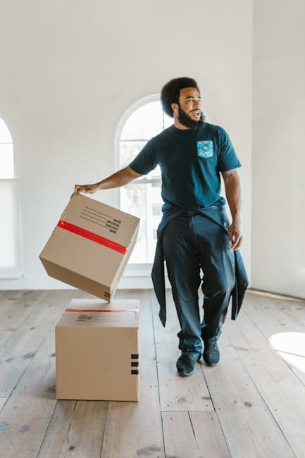 A person in casual clothing, wearing a wristwatch and a bracelet, is captured during the home relocation process as they use a tape measure on a large cardboard box inside a well-lit room with large w