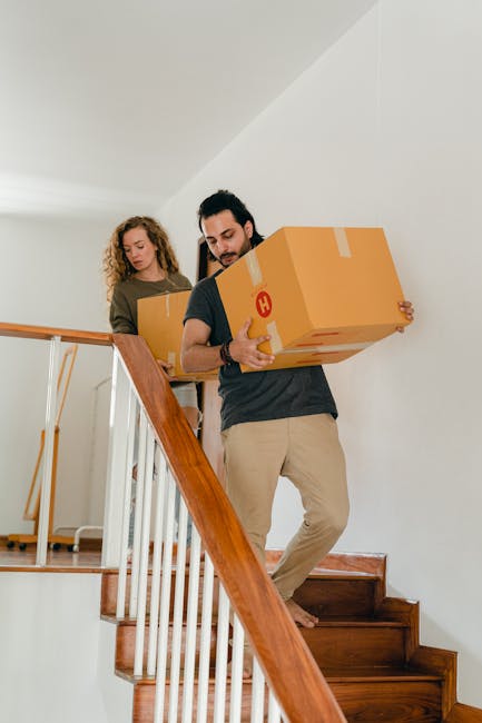 A man and a woman are engaged in a home relocation inside a property, moving cardboard boxes on a staircase with wooden steps and white risers. The man, positioned at the forefront, is carrying a large cardboard box with a red and white label, holding it securely with both hands as he descends the staircase. He is dressed in a dark t-shirt and light-colored trousers. Behind him, the woman has curly hair and is also carrying a cardboard box, walking down the stairs carefully. The staircase has a wooden handrail on the left side, and the interior features white walls and a simple, clean decor. The lighting appears natural, illuminating the space evenly. This scene depicts the loading process involved in a furniture transport or packing and moving service, with a focus on careful handling of boxes during the home relocation process, as provided by Man with Van Homerton.