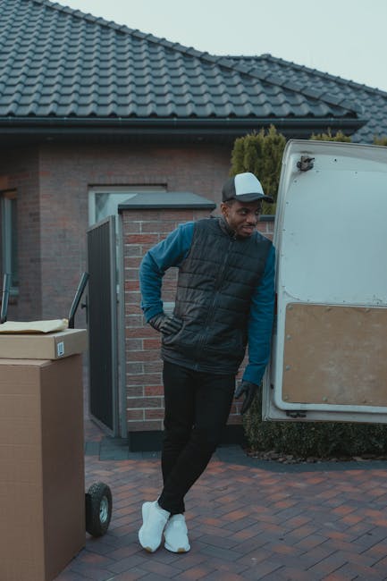 A man in a blue long-sleeve shirt wearing a black quilted vest, black pants, white sneakers, and a grey cap stands beside an open white moving van parked on a paved driveway outside a residential prop
