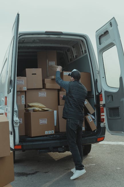 A man in a blue long-sleeve shirt wearing a black quilted vest, black pants, white sneakers, and a grey cap stands beside an open white moving van parked on a paved driveway outside a residential prop
